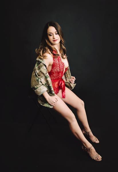 Woman in red lingerie and camo jacket, seated with a black background.