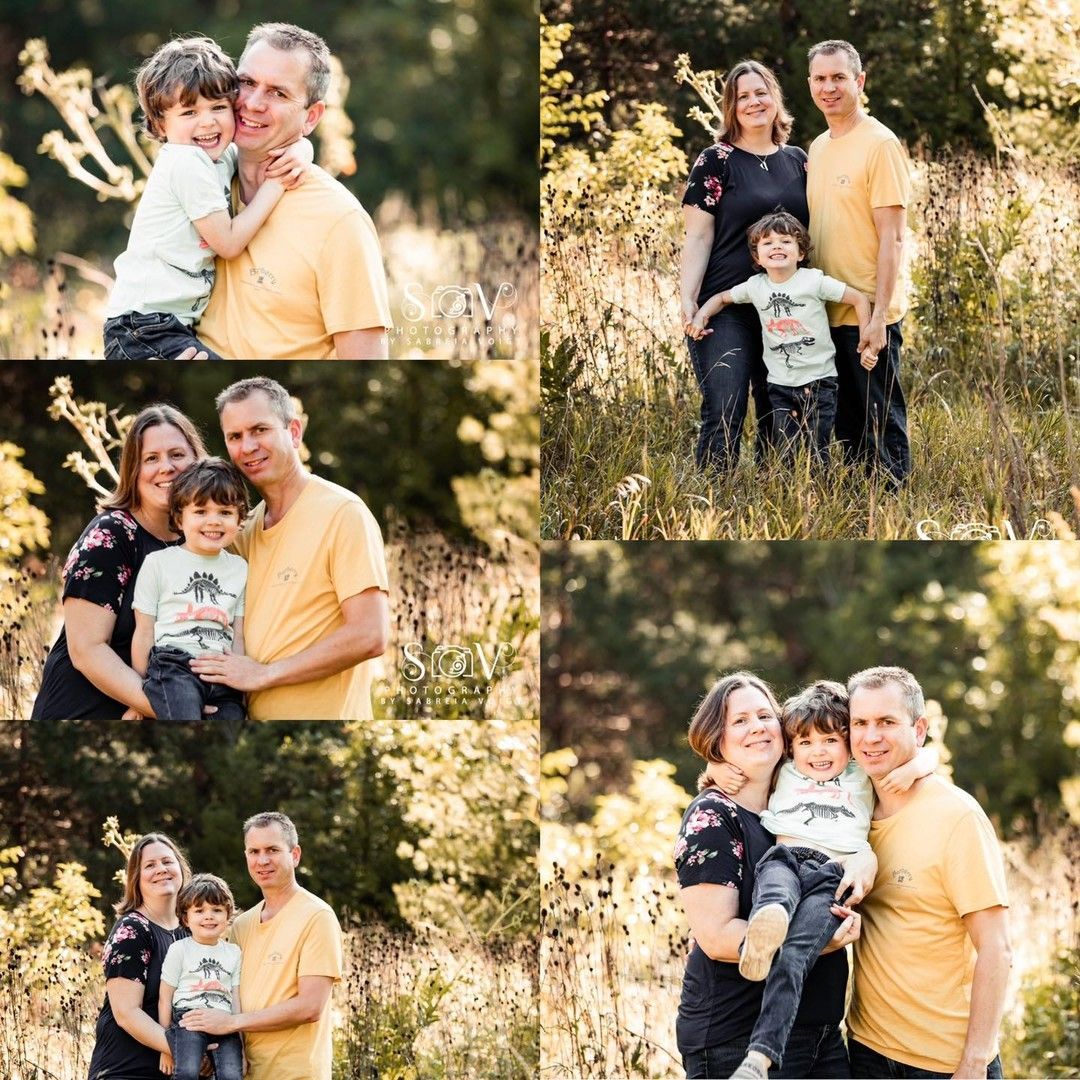 Family poses outdoors in tall grass; father in yellow shirt, mother in black, child in graphic tee.