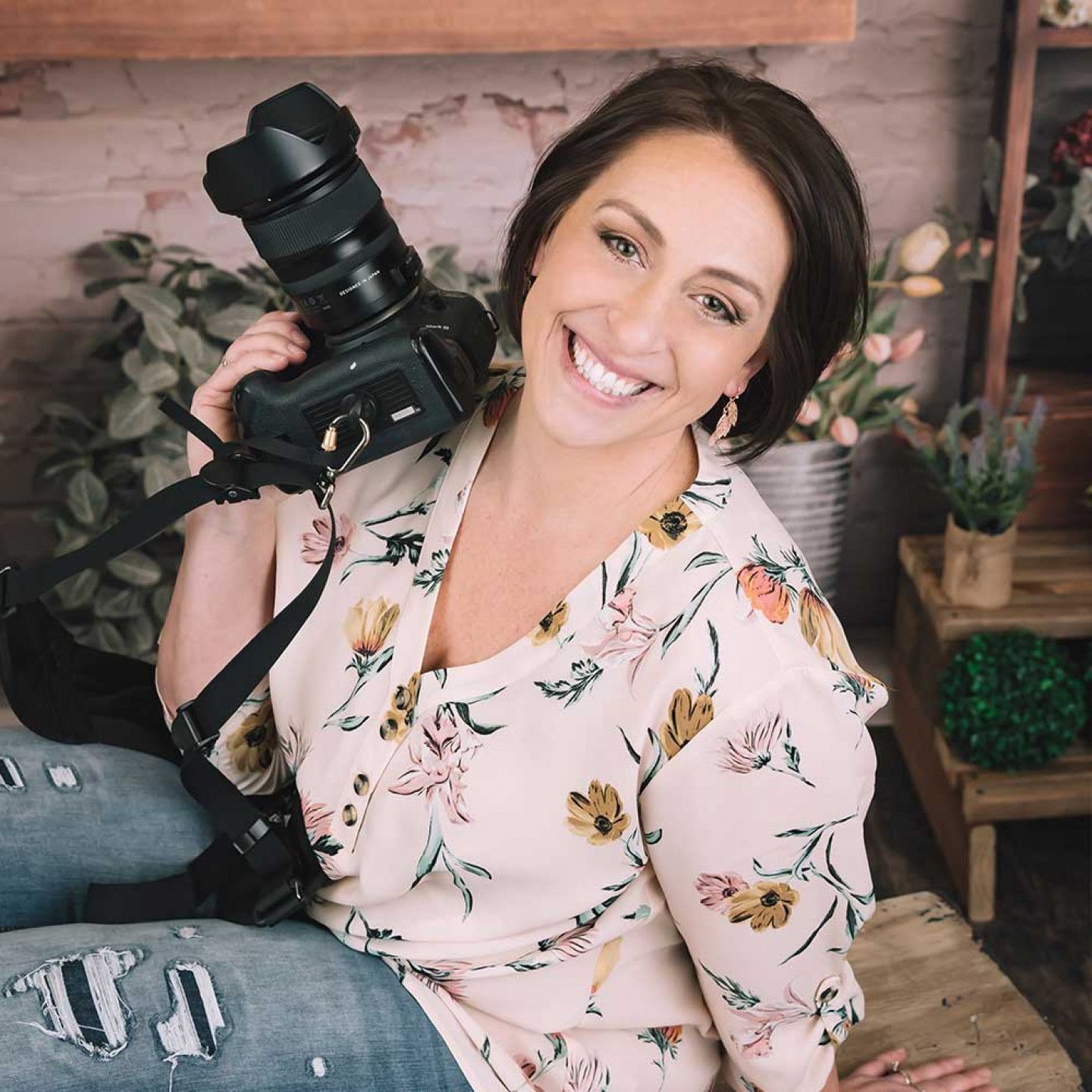 Woman smiling, holding a camera, wearing floral shirt, sitting near flowers and greenery.