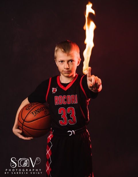 Boy in basketball uniform holds ball and points to finger with a flame against a dark background.