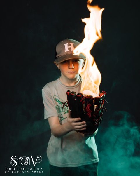 Boy holding flaming baseball glove against a smoky, dark background; wearing a baseball cap and t-shirt.