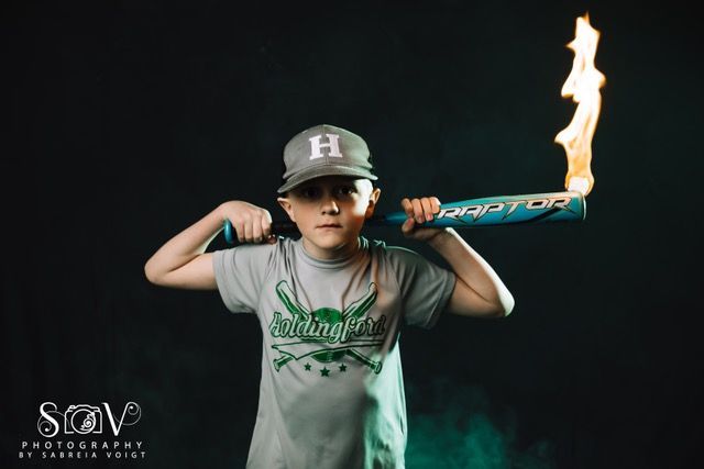Boy with a baseball bat on fire, wearing a hat and shirt, against a dark background.