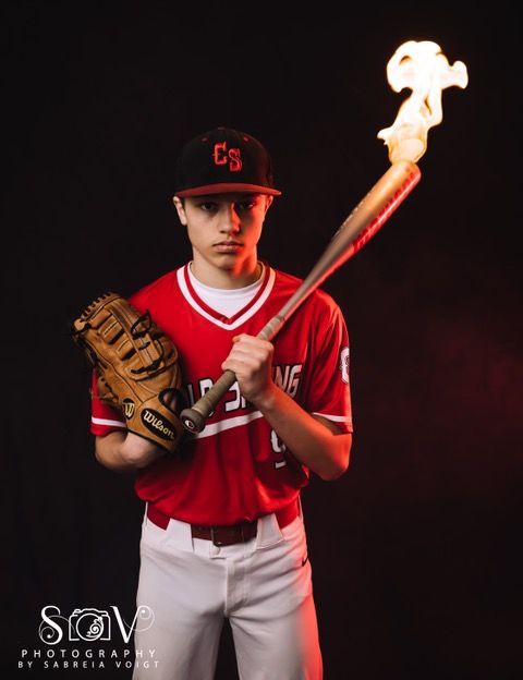 Baseball player holding burning bat and glove, red uniform, black background.