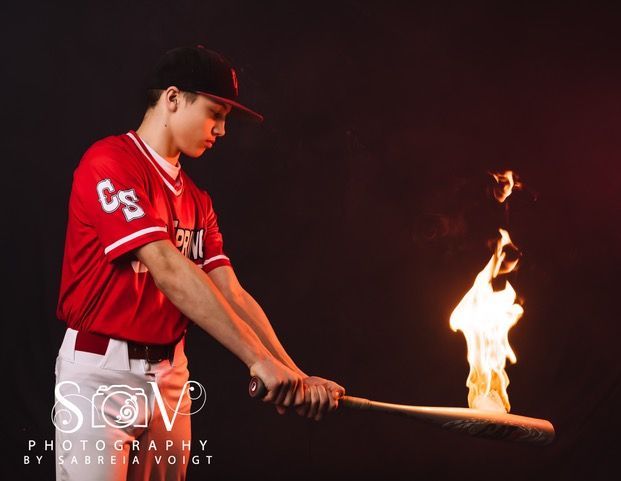 Baseball player in red uniform holds flaming bat against a dark background.