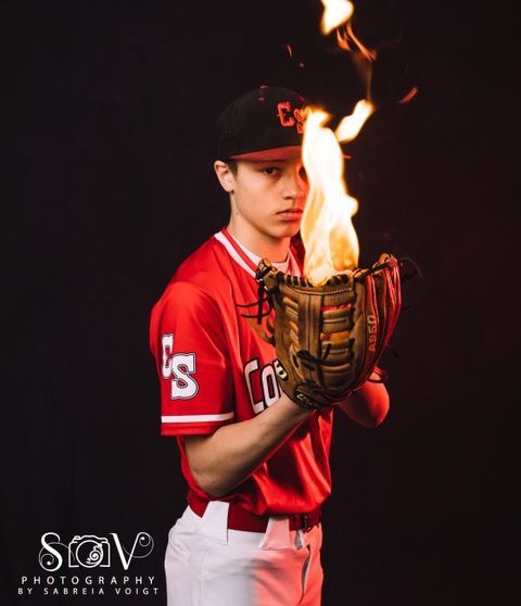 Baseball player holding a mitt with flames, red jersey, black cap, dark background.