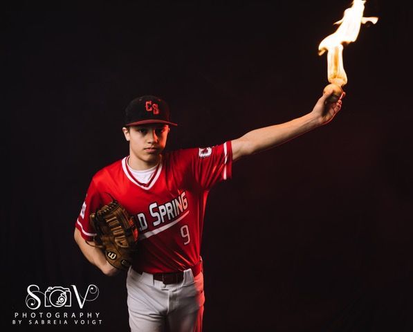 Baseball player in red uniform with flaming ball, black background.