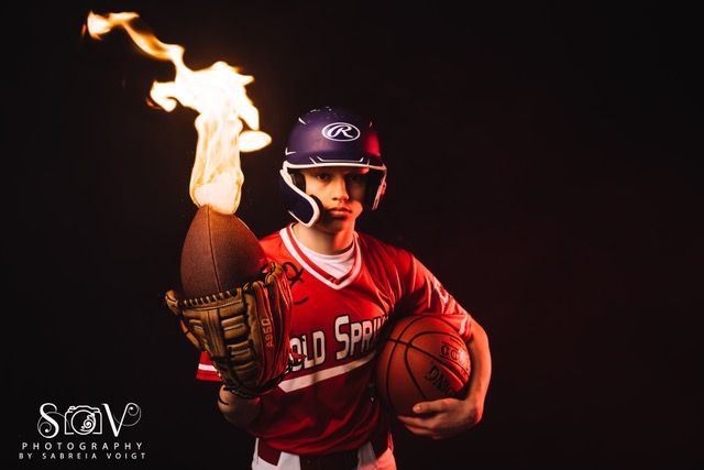 Baseball player holding flaming football and basketball, wearing helmet and uniform. Dark background.