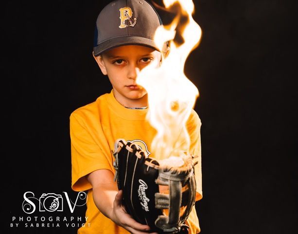 Boy in baseball cap holds burning baseball glove against black backdrop.