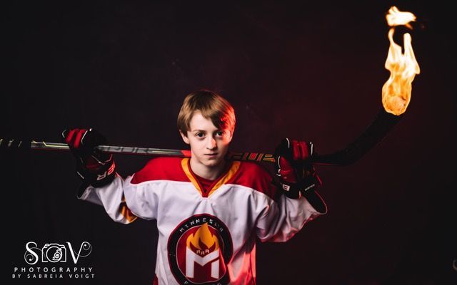 Young hockey player in red and white jersey holds flaming hockey stick over his shoulders, dark background.