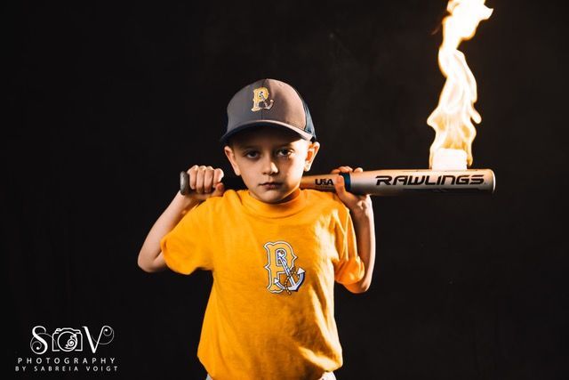 Young baseball player holding a flaming bat, wearing a yellow shirt and a baseball cap against a black background.