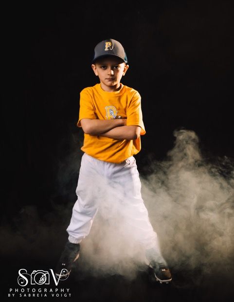 Boy in baseball uniform stands confidently, arms crossed, amidst fog against a black backdrop.