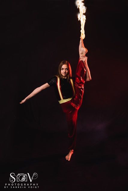 A dancer in a vertical split, holding fire, against a dark background. Red and black clothing.