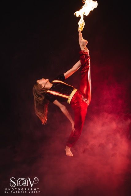 Person in red pants and top performs a vertical split, foot holding a flaming torch against a red-tinted backdrop.