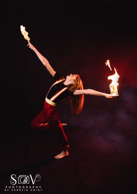 A person balances, holding flames aloft. They wear red pants, a black top, and stand against a dark backdrop.