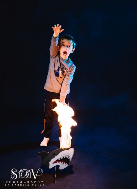 Child with open mouth, reaching up. A toy shark with flames coming from its mouth, set against a dark blue background.