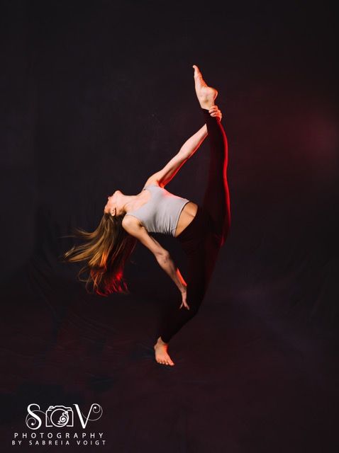 Dancer in mid-air, leg extended upward. She wears a gray top, black leggings, and performs against a dark red lit background.