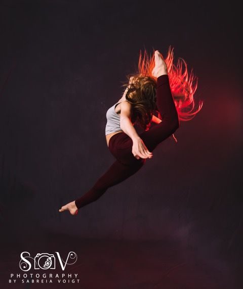 Dancer in mid-air split, hair flowing, wearing a gray top and maroon leggings, against a dark red backdrop.