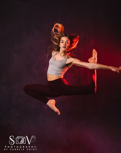 A dancer in mid-air, leg extended. Grey top, burgundy pants. Dramatic red and purple backdrop.
