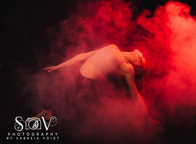 A dancer floats in red smoke, arms outstretched, head tilted back. Dark background.