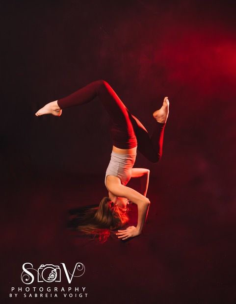 A dancer performs a handstand with legs curved, lit by red light on a dark background.
