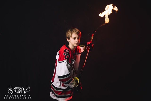 Boy in hockey jersey holds flaming hockey stick, red and white, dark background.