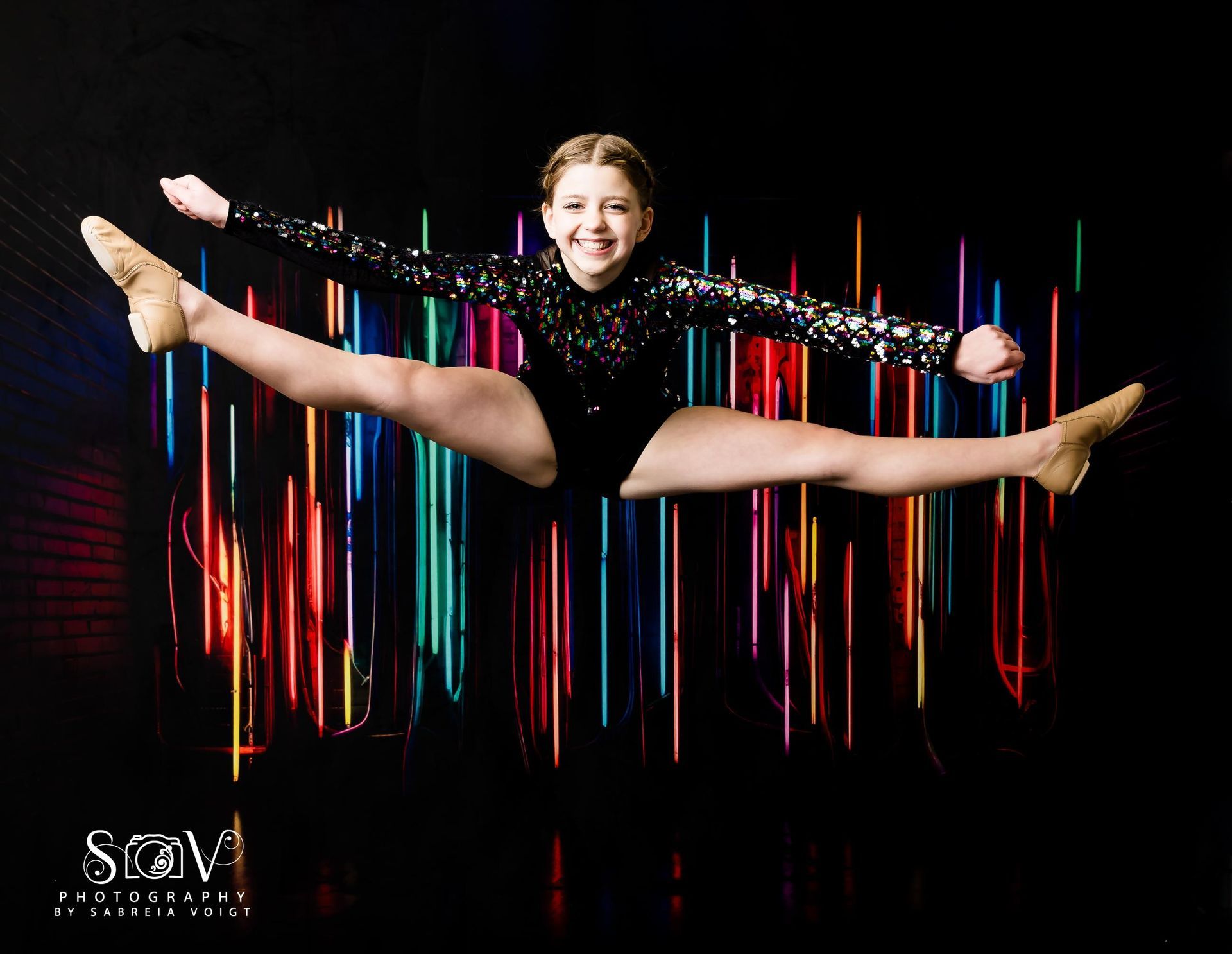 Dancer in black sequin outfit performs a split leap against colorful light trails on a dark backdrop.
