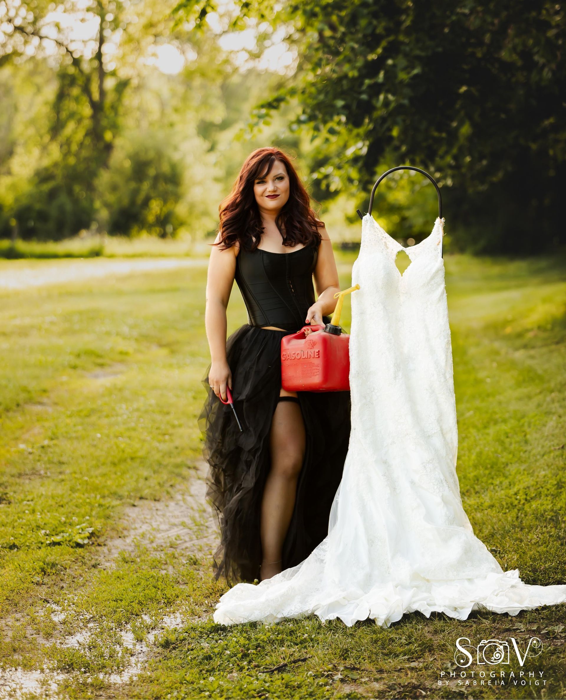 Woman in black corset and tulle skirt, holding gas can, beside a white wedding dress in a grassy field.