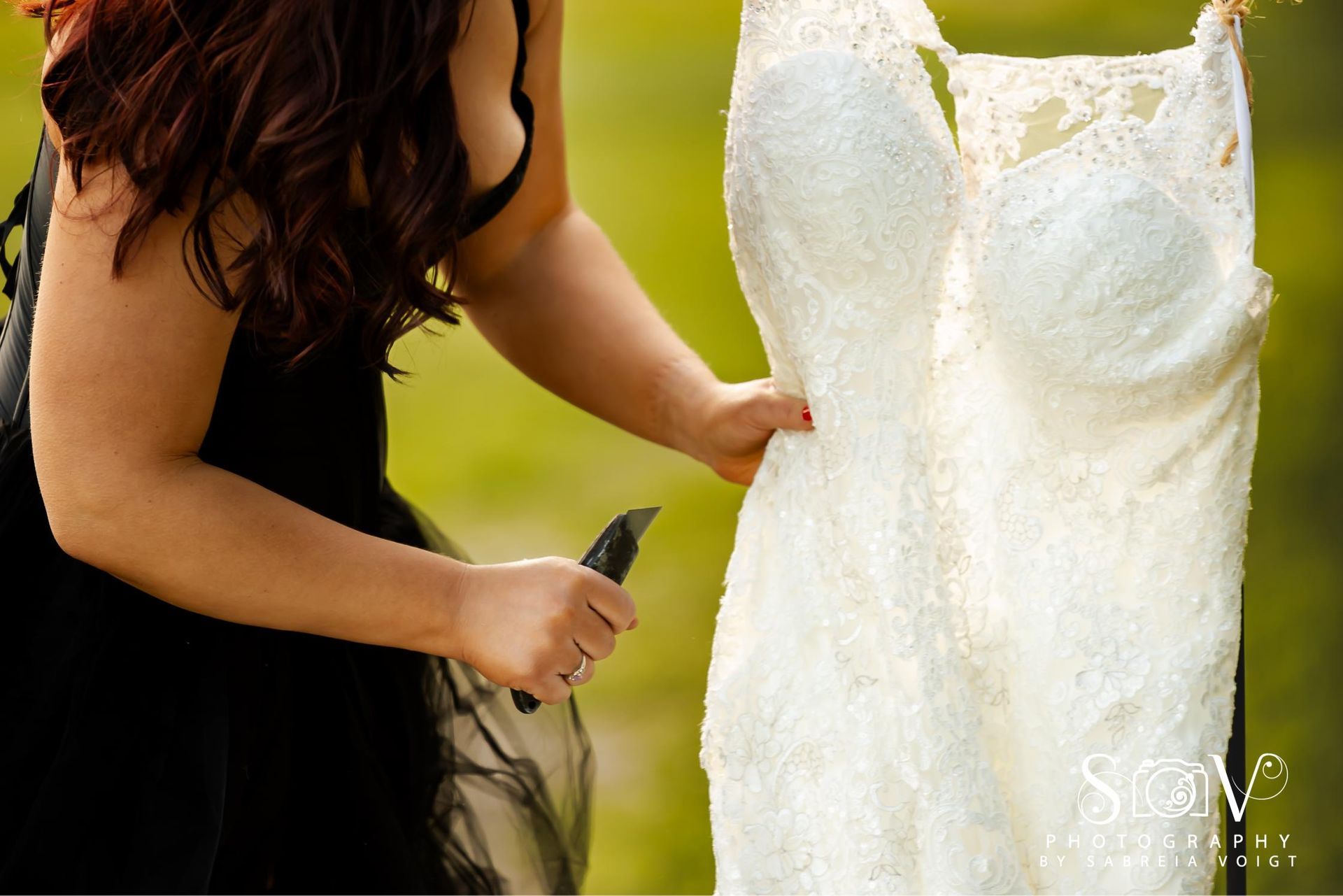 Woman in black dress holding a knife near a hanging white wedding dress. Outdoor setting.