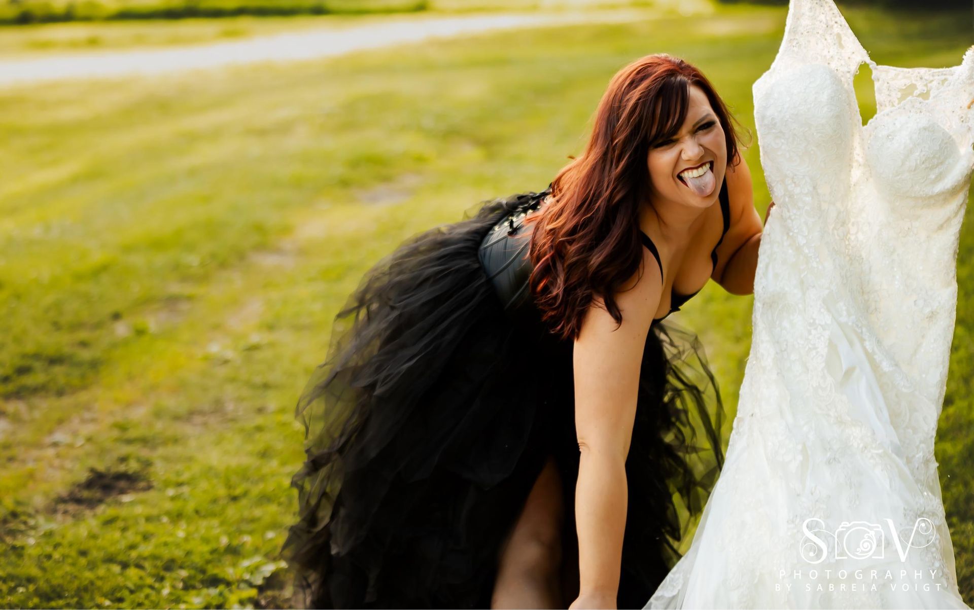 Woman in black tutu sticking out tongue next to a white wedding dress in a grassy field.