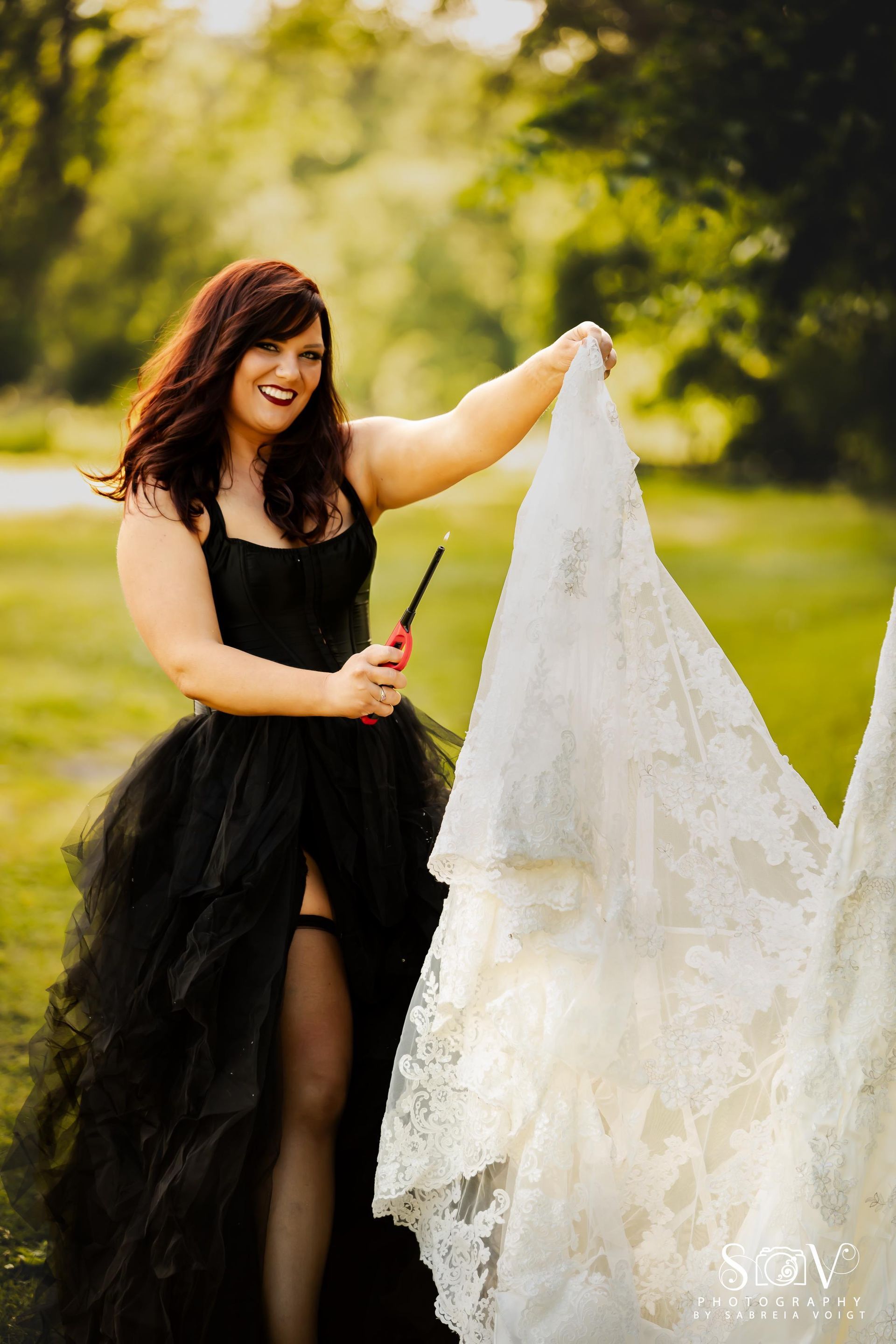 Woman in black dress, holding white wedding dress, about to light it with a lighter in an outdoor setting.