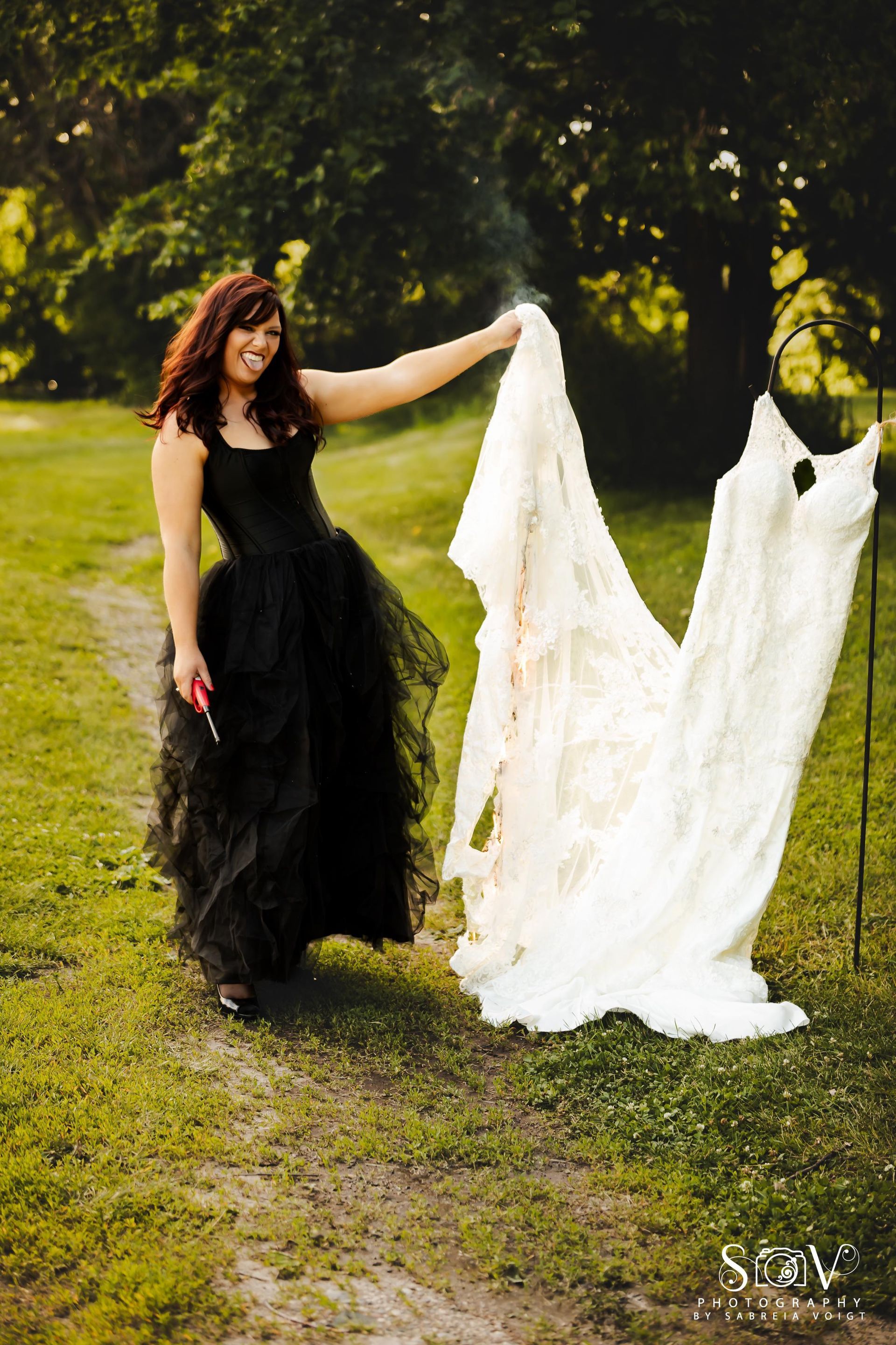 Woman in black dress holds up a white wedding gown outdoors.