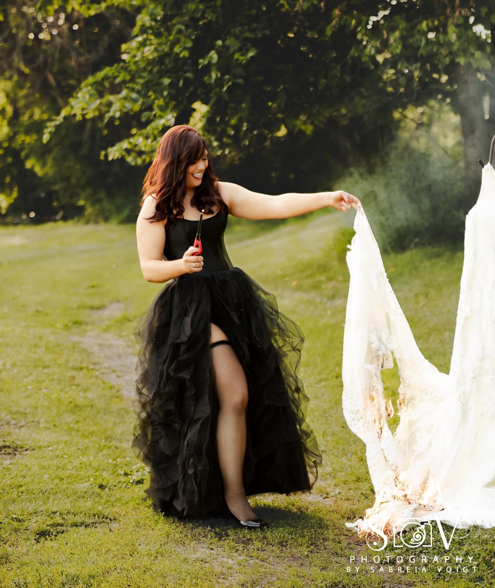 Woman in black dress pops bubbles, holding a tattered white wedding dress in grassy area.