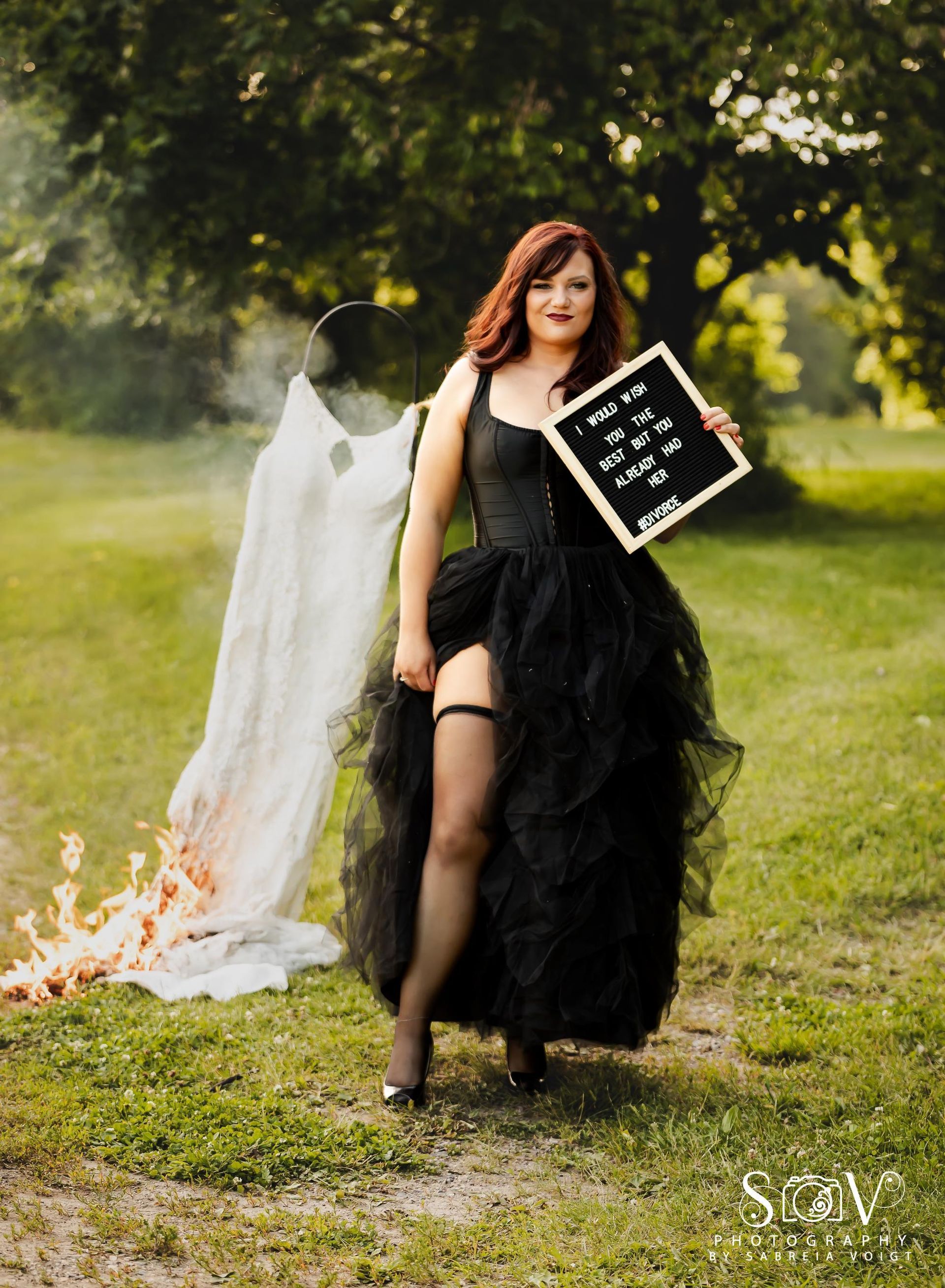 Woman in black dress and garter, holding sign. White cloth and fire, with a tree in the background.