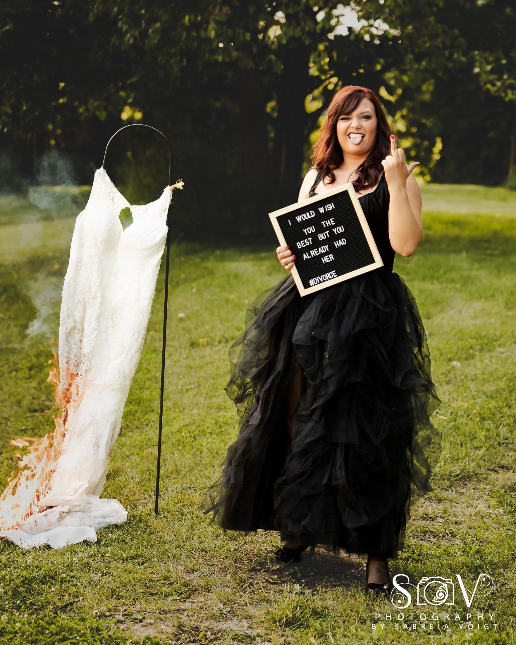 Woman in black gown holds sign, flipping off burning wedding dress outdoors.