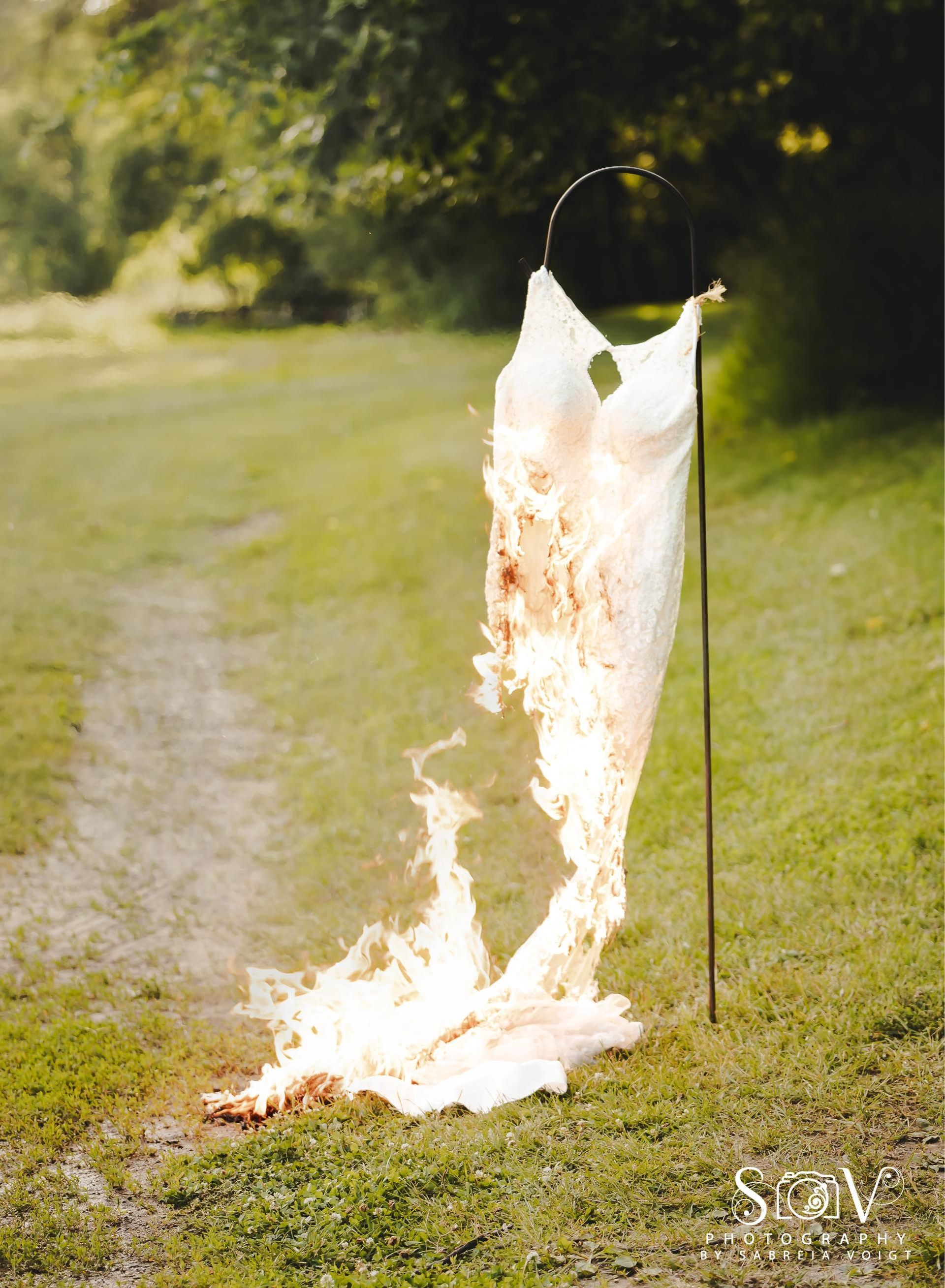 Burning white wedding dress on a metal pole in a grassy field. Flames engulf the dress.