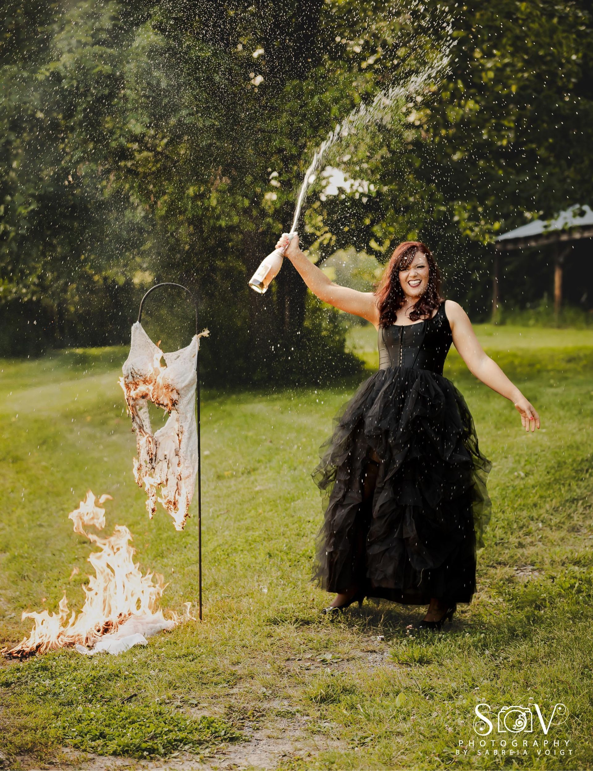 Woman in black gown celebrating with champagne, next to a burning object outdoors.