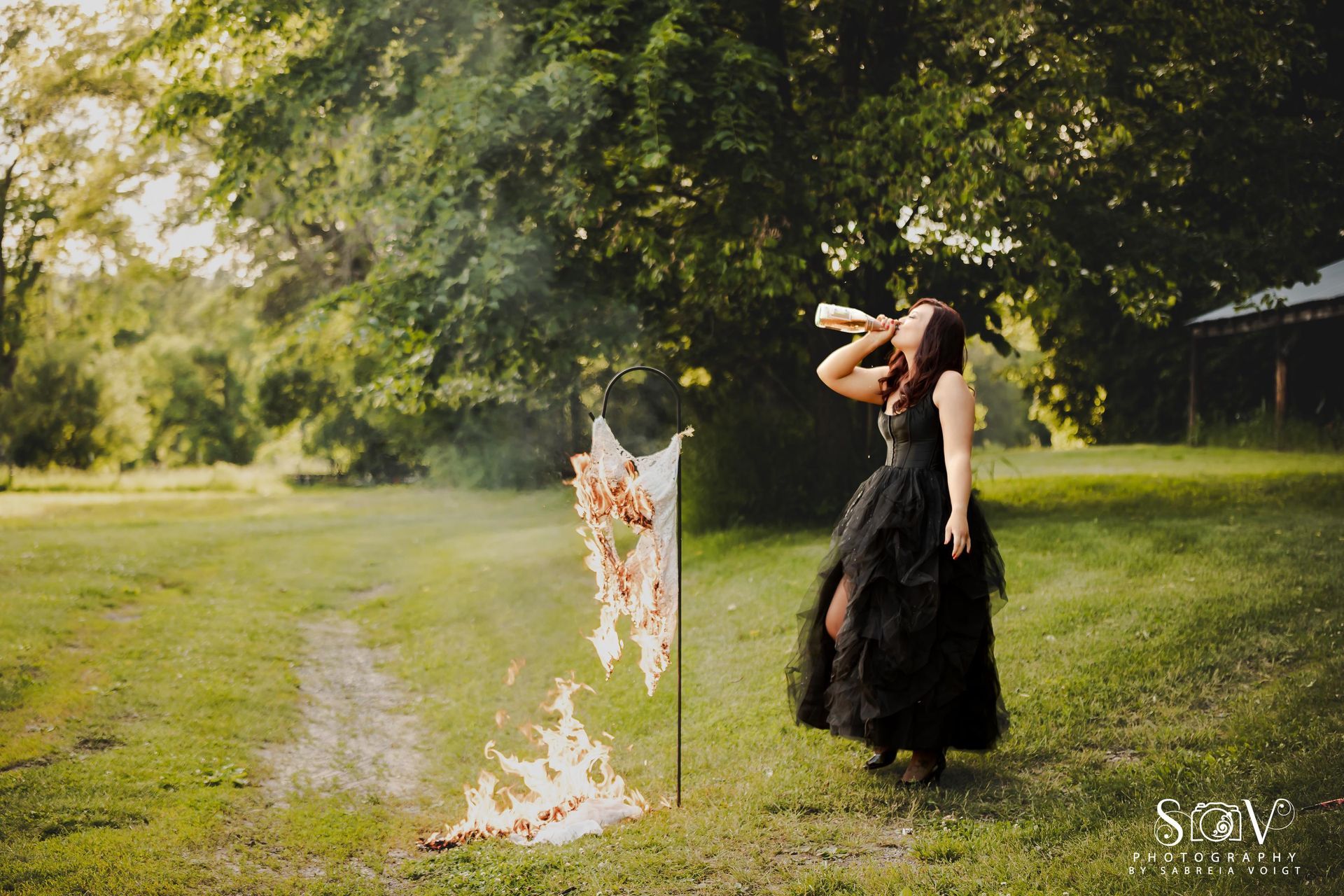 Woman in black dress drinking from a horn, next to burning sign in grassy field.