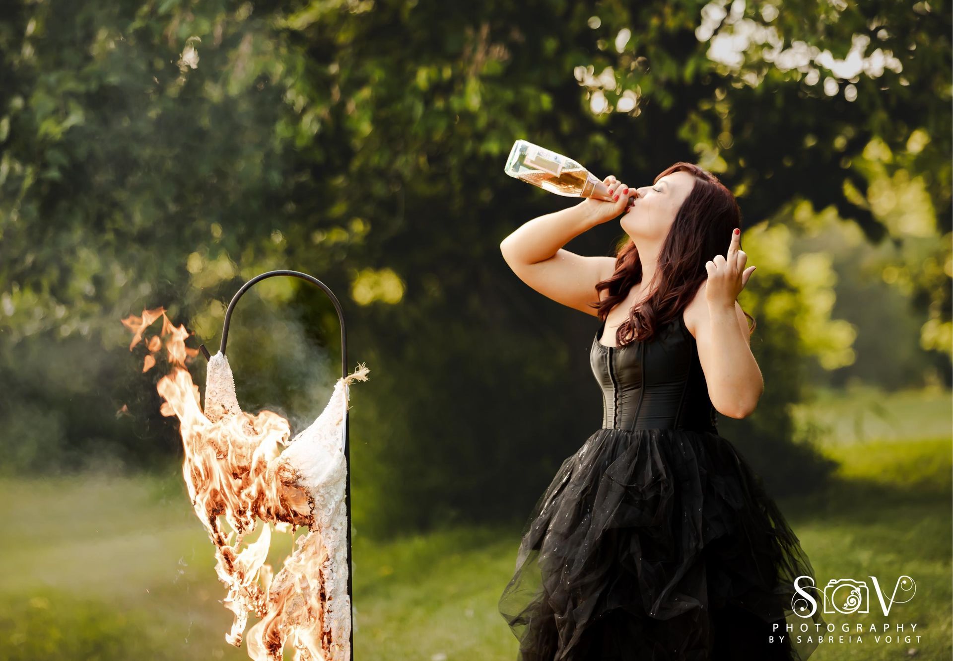Woman in black dress drinking from a bottle, burning effigy in the background, outdoor setting.