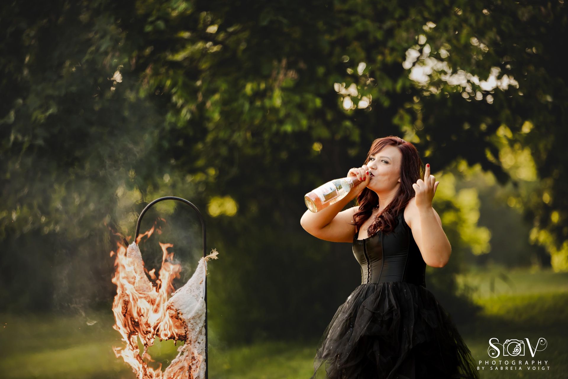 Woman in black dress drinking, burning wedding dress outdoors.
