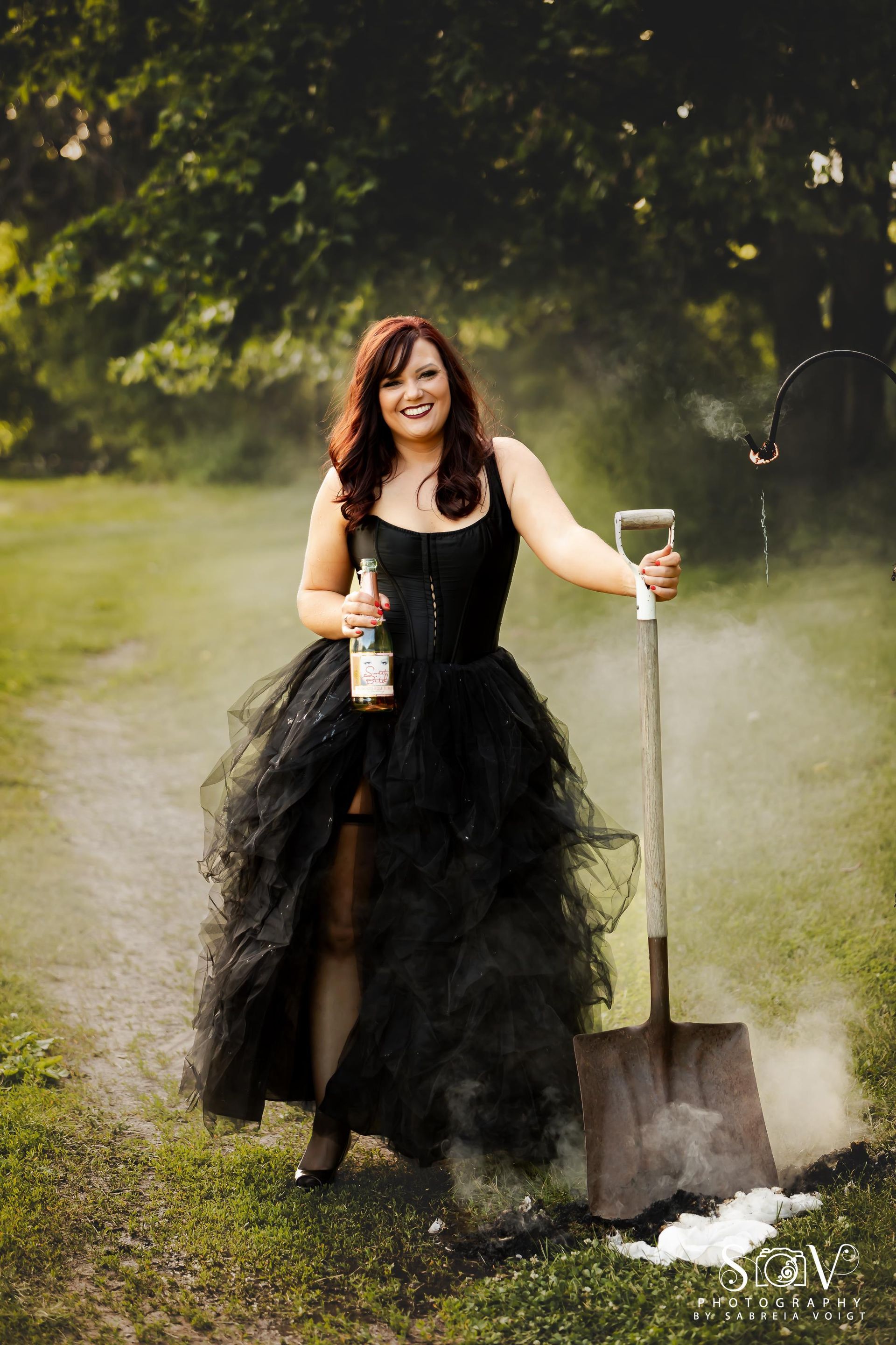 Woman in black dress with champagne and shovel in a field, smoke around the shovel.