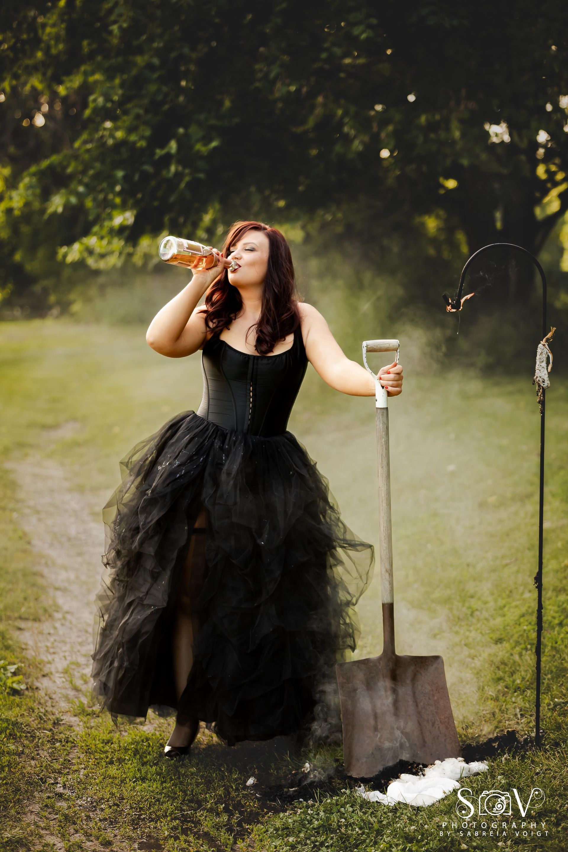 Woman in black dress drinking from a bottle, leaning on a shovel in a field.