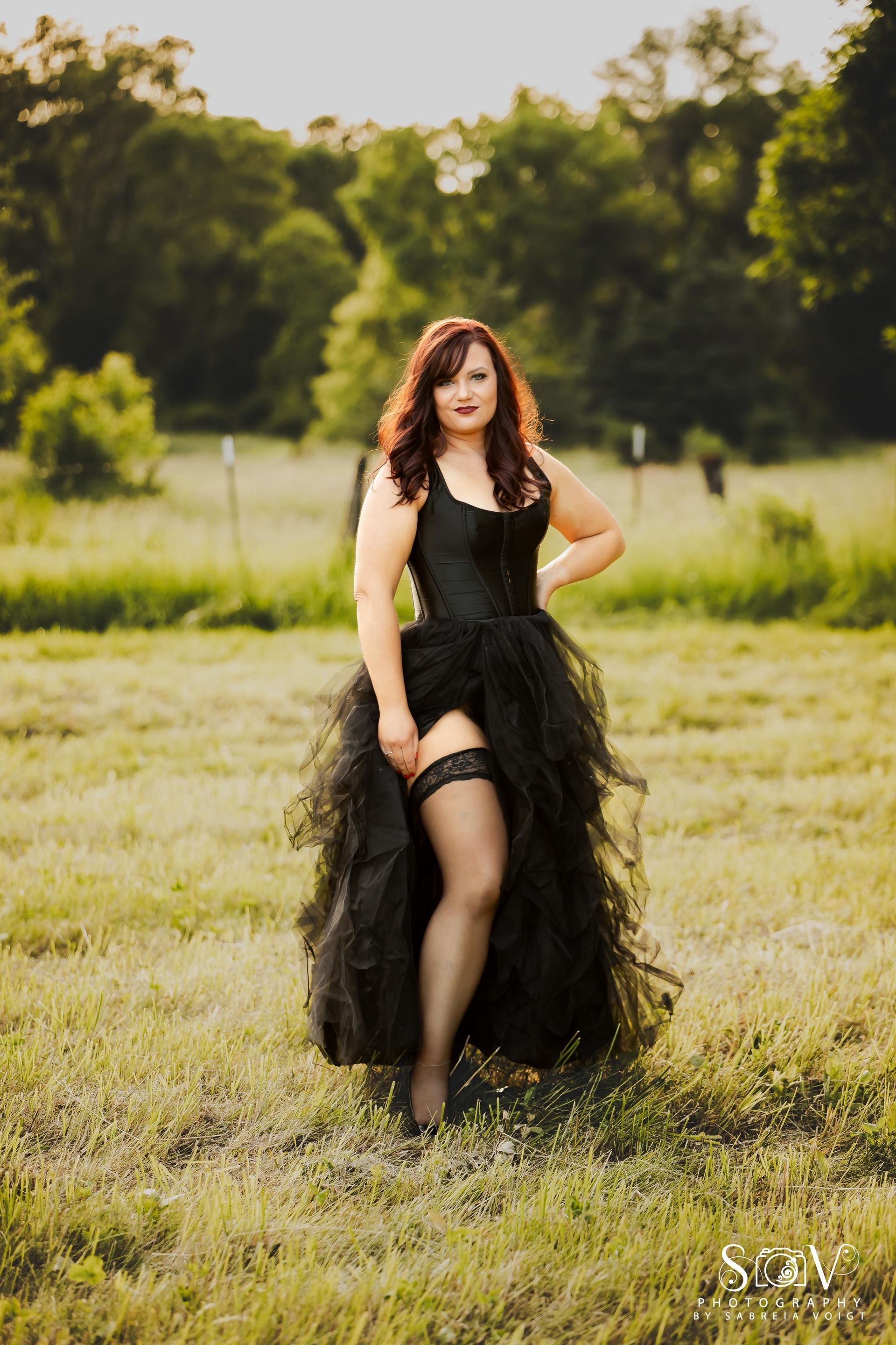 Woman in black corset and tulle skirt, thigh high stocking, posing in grassy field.