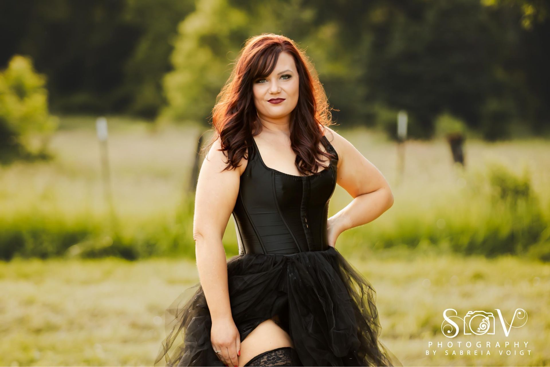 Woman in black corset and tulle skirt poses outdoors in a field, hand on hip.