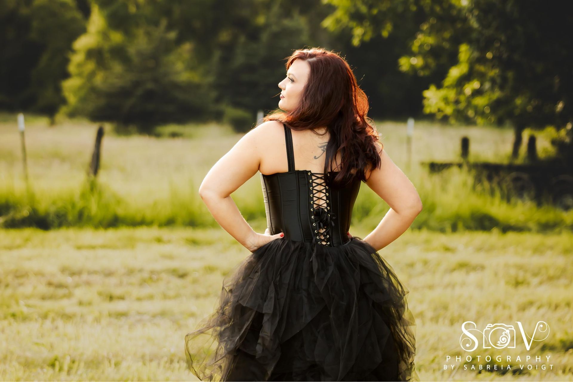 Woman in black corset and tulle skirt stands in a field, hands on hips, looking right.