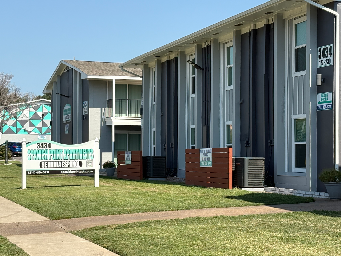 Apartment building with gray and white exterior, sign on lawn, sunny day.