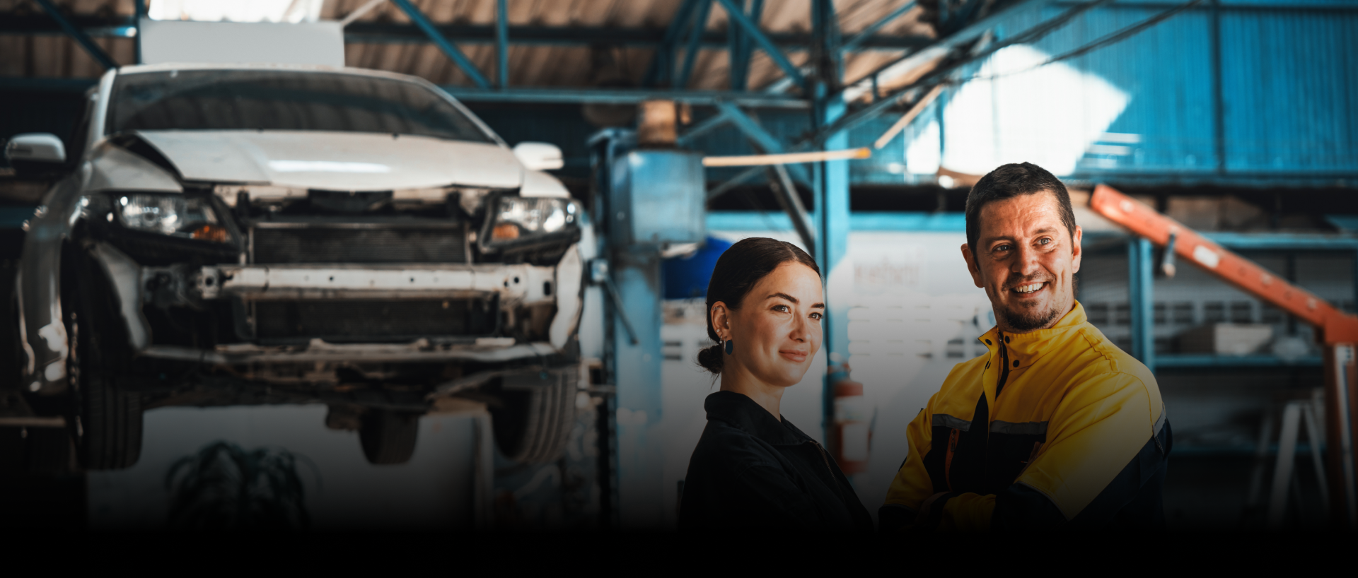 A mechanic and a woman smiling in a car repair shop with a car on a lift. | Strong's Alignment Service
