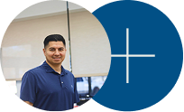 Man in a navy polo standing beside a blue circle with a white plus sign in an office setting