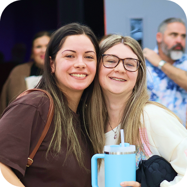 Two smiling friends posing indoors, one holding a blue tumbler