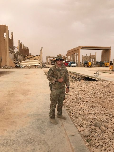 Cole Gasperini in uniform standing near partially destroyed buildings in a dusty, outdoor setting.