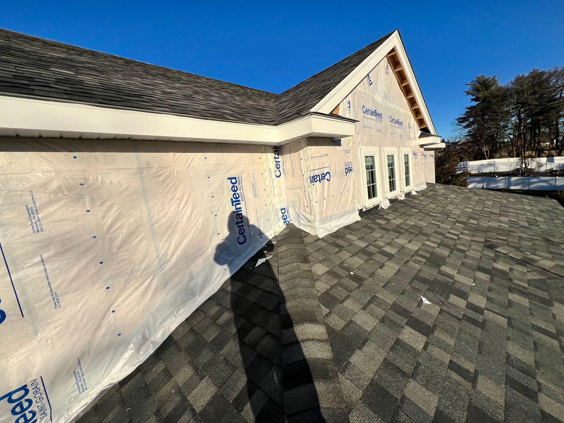 Roof of a house under construction with dark gray shingles, white trim, and blue wrap on walls against a clear sky.